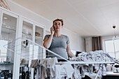 Pregnant woman talking on phone while doing housework and hanging baby clothes on drying rack