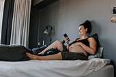 Happy pregnant woman relaxing in bed and using phone