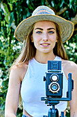 Porträt einer jungen schönen kaukasischen Frau in den 20ern mit blauen Augen, die einen Hut trägt und mit einer alten Vintage-Kamera auf einem Stativ im Freien in einem Garten fotografiert. Lifestyle-Konzept.