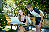 Portrait of a young caucasian woman with her younger brother posing outdoor in a garden with a laptop looking information in internet. Lifestyle concept.