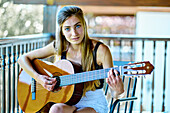 Porträt einer jungen schönen kaukasischen Frau in den 20ern mit langen Haaren und blauen Augen, die auf der Veranda eines Landhauses Gitarre spielt. Lebensstil-Konzept.