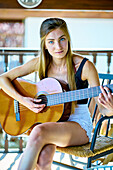 Portrait einer jungen schönen kaukasischen Frau in den 20ern mit langen Haaren und blauen Augen, die auf der Veranda eines Landhauses Gitarre spielt. Lifestyle-Konzept.