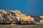 Eine Reihe von Eisenbahnwaggons mit Mittelträgern in der Green River Wüste von Utah mit Scheinwerfern auf den Book Cliffs dahinter.