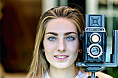 Portrait einer jungen schönen kaukasischen Frau in den 20ern mit blauen Augen, die mit einer alten Vintage-Kamera auf einem Stativ im Freien fotografiert. Lifestyle-Konzept.