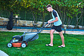 Portrait of a young caucasian boy passing the mower through in the garden in a country house. Lifestyle concept.