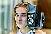 Portrait einer jungen schönen kaukasischen Frau in den 20ern mit blauen Augen beim Fotografieren mit einer alten Vintage-Kamera auf einem Stativ im Freien. Lifestyle-Konzept.