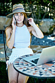 Porträt einer jungen schönen kaukasischen Frau in den 20ern mit Hut und im Freien in einem Garten, die mit ihrem Handy telefoniert und mit einem Laptop nach Informationen im Internet sucht. Lebensstil-Konzept.