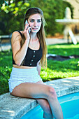 Porträt einer jungen schönen kaukasischen Frau in den 20ern mit langen Haaren und blauen Augen, die im Freien am Rande eines Swimmingpools sitzt und mit ihrem Handy telefoniert. Lifestyle-Konzept.