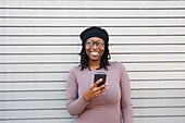 Lächelnde Frau mit Brille und Baskenmütze hält ein Smartphone in der Stadt