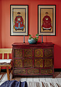 Antique painted wooden chest of drawers with Asian flair in front of orange wall with framed pictures