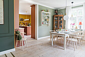 Dining area with light-colored wooden furniture and green walls with view into kitchen