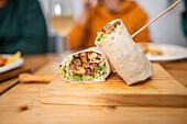 Closeup of delicious tortilla roll sliced and wrapped in lettuce and vegetables on wooden table against blurred background of crop people eating over table