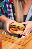Crop blond female in casual clothes sitting at wooden table and eating delicious burger with glass of beverage in cafe