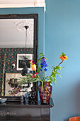 Buntes Blumenarrangement in roter Glasvase auf schwarzem Kaminsims vor blau gestrichener Wand