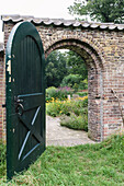 Open green wooden gate leads into a flowering garden with stone archway