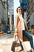 Junge Frau mit Brille und Tasche in hellem Mantel auf der Straße mit Hochhäusern