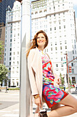 Young woman with glasses in a colorful dress and a light coat stands on the street