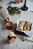 Life style scene. Cookies with mint leaves in a wooden box on linen table close to Advent gift box. Woman s hand in a wool jamper holds a red cup of coffee