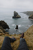 POV Beine eines Mannes, der auf einer Klippe an der zerklüfteten Küste sitzt, Duncansby, Schottische Highlands, Schottland