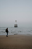 Woman walking on ocean beach with lighthouse, Rattray, Aberdeenshire, Scotland\n