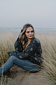 Portrait beautiful young woman sitting among beach grass, Rattray, Aberdeenshire, Scotland\n
