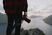 Fotograf hält Spiegelreflexkamera auf einem Hügel über dem Meer, Klakkur, Klaksvik, Färöer Inseln