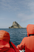 Touristen auf einer Fähre mit Blick auf eine Felsformation über dem sonnigen Meer, Mykines, Färöer Inseln