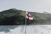 Flagge der Färöer Inseln auf Fährboot auf sonnigem Meer unterhalb der Klippen, Mykines, Färöer Inseln