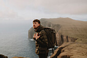 Mann mit Rucksack beim Wandern auf Klippen über dem Meer, Asmundarstakkur, Suduroy, Färöer Inseln