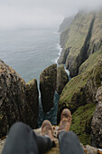 POV hiker sitting on cliff above sea stacks over ocean, Dunnesdrangar, Vagar, Faroe Islands\n