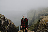 Hiker in fog on cliffs above sea stacks, Dunnesdrangar, Vagar, Faroe Islands\n