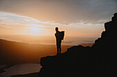 Silhouetted hiker standing on mountain, looking at dramatic sunset sky, Assynt, Sutherland, Scotland\n