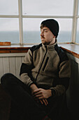 Thoughtful male hiker in beanie sitting at window, looking away\n