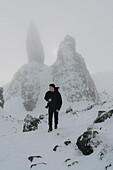 Male hiker with camera hiking below snow covered rock formation, Old Man of Storr, Scotland\n