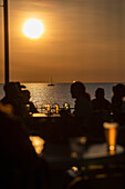Silhouetted people drinking beer on waterfront patio with ocean view at sunset\n