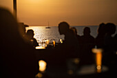 Silhouetted people drinking beer on ocean waterfront patio at sunset\n