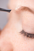 Extreme close up of young woman plucking own eyebrow hair with tweezers\n