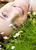 Junge Frau liegt auf einer Blumenwiese mit Gänseblümchen im Mund