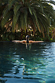 Back of young woman standing in an exotic swimming pool with arms stretched\n