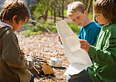 Three boys kneeling around campfire looking at map\n