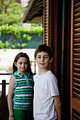 Young boy and young girl standing by window with wooden shutters\n