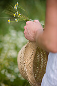 Close up of a woman hand holding a straw hat and camomile flowers\n