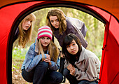Teenage girls at campsite crouching in front of tent entrance looking in with curiosity\n