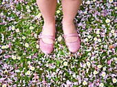 Junges Mädchen mit Füßen und Beinen in rosa Schuhen steht auf einem mit Blütenblättern und Blumen bedeckten Rasen