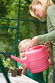 Young blonde girl helping woman watering seedling pots in the greenhouse with pink watering can\n