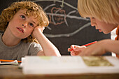 Two young boys writing and sitting at desk in the classroom one of them is day dreaming\n