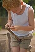 Young boy holding wooden stick between legs tying a knot with a string\n