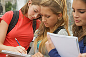 Three teenaged girls reading, writing and holding note pads\n