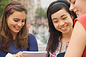 Three smiling teenaged girls reading, writing and holding note pads\n