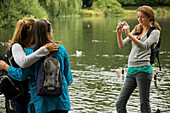 Teenaged girl taking photograph of two women by a lake\n
